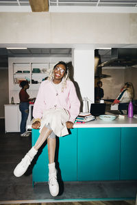 Portrait of smiling young woman sitting on dining table in kitchen at college dorm