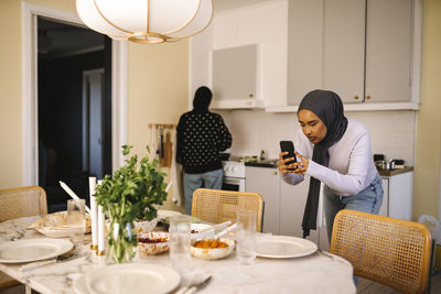 Young woman photographing food through mobile phone while friend cooking in kitchen at home
