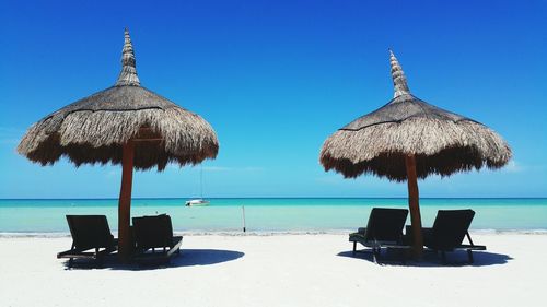 Thatched roofs over lounge chairs at beach against blue sky