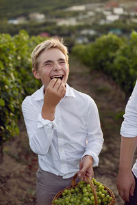 Teenage boy in a white shirt stands in a vineyard at sunset and holds a basket of green grapes