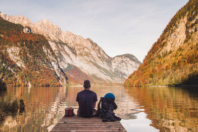 Rear view of man sitting on pier over lake against mountains