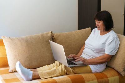 Elderly woman smiling 60s, sitting at home on couch, holding laptop. senior