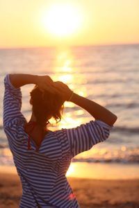 Rear view of woman with hands behind head standing at beach during sunset