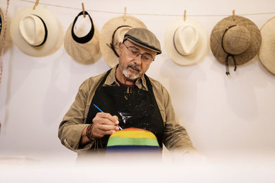 Elderly male master in apron painting rainbow pattern on stylish straw hat with brush standing at workbench with colorful dyes