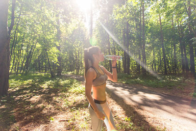 Young woman standing by trees in forest on sunny day