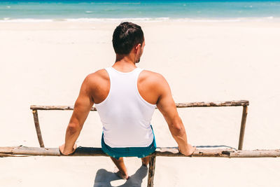 Rear view of man sitting on parallel bars at beach during sunny day