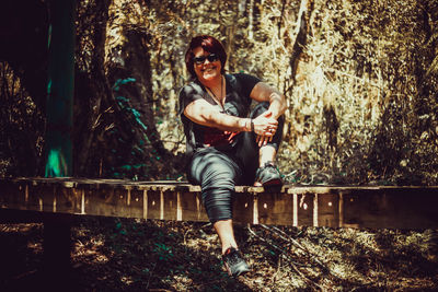 Portrait of smiling woman wearing sunglasses sitting on footbridge in forest