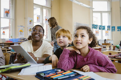 Smiling teacher holding tablet pc sitting with schoolboy and schoolgirl at desk while looking up in classroom