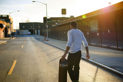 Rear view of man carrying skateboard while walking on road in city