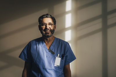 Portrait of happy male doctor standing against wall at hospital