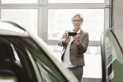 Senior businesswoman photographing car through smart phone in dealership store