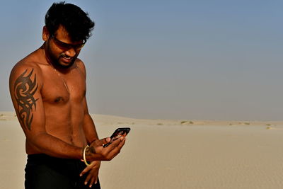 Shirtless young man taking selfie with mobile phone at desert during sunny day