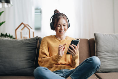 Smiling woman listening music through wireless headphones while using smart phone at home