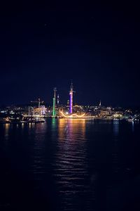 Illuminated buildings by sea against sky at night