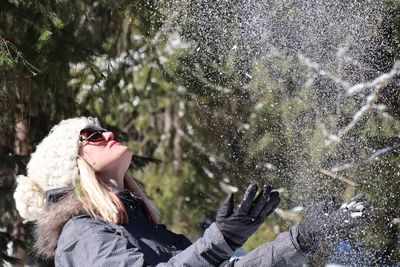 Side view of playful woman throwing snow in forest during winter