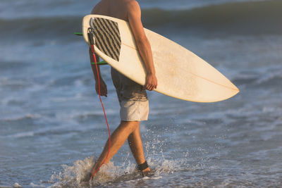Low section of man holding surfboard while walking on shore at beach