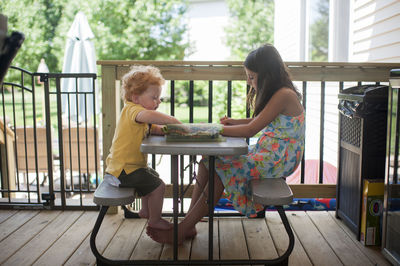 Side view of siblings with colorful crayons in box sitting on bench at porch