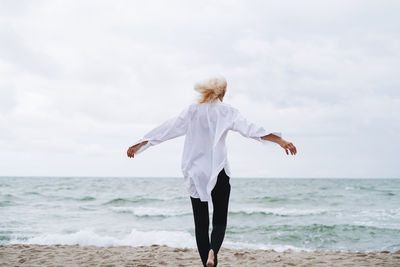 Portrait of elegant blonde woman in white shirt on sand beach at storm sea at windy weater