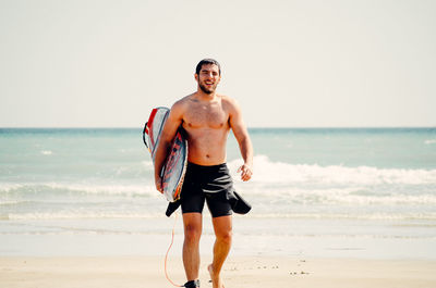 Young man with surfboard standing on beach against sky