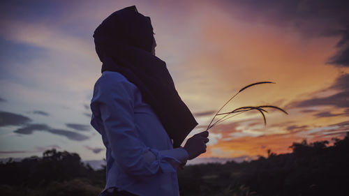 Side view of woman holding plant against sky during sunset