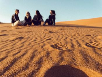 Friends sitting on sand at beach against clear sky