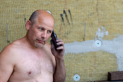 Shirtless mature man using smart phone while standing against wall