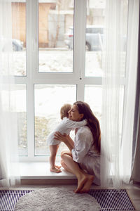 Mother in a white robe sits with a child a blonde daughter at a large window of the house person