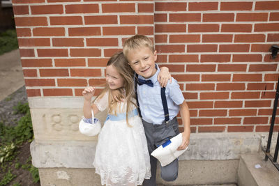 Flower girl and ring bearer stand against brick wall hugging