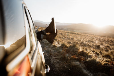 Unrecognizable tourist lying in car with legs out of window on background of sunrise in scottish highlands