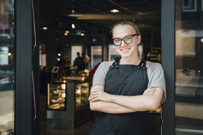 Portrait of smiling andorgynous owner standing with arms crossed at cafe doorway