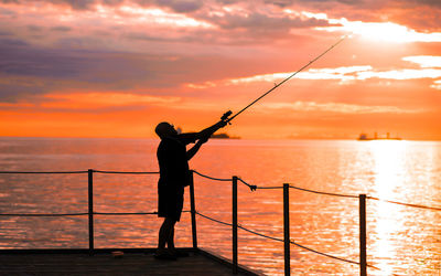 Silhouette man fishing by sea against sky during sunset