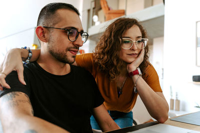 Side view of young bearded ethnic man and curly haired woman in casual clothes and eyeglasses working together on laptop during remote job at home