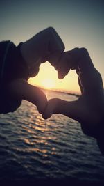 Close-up of hands making heart shape against sea during sunset