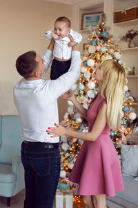 Dad raises his son in his arms standing next to the christmas tree in the room