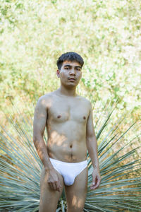 Portrait of shirtless man wearing underwear standing against tree