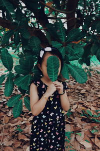 Girl covering face with leaf while standing against tree