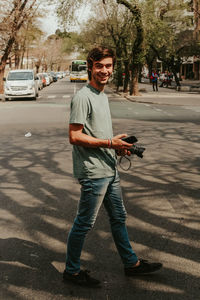 Full length portrait of young man holding camera on road