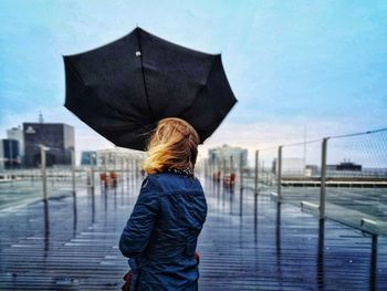 Rear view of woman holding umbrella while standing by railing against sky