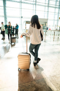 Back view of unrecognizable female traveler in casual clothes with dark hair leaning on suitcase while standing in sunlit terminal of modern airport