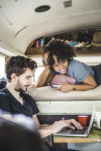 Smiling young woman lying on bed while looking at man using laptop in motor home