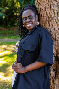 Portrait of a smiling young woman standing against tree trunk
