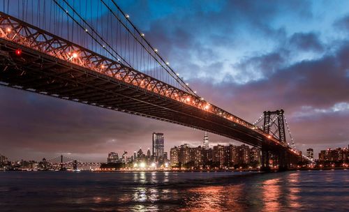 Illuminated suspension bridge over river against cloudy sky