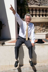 Full length of businessman pointing while sitting on retaining wall on sunny day