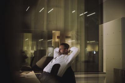 Mature businessman with hands behind head sitting in office at night