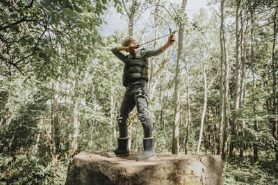 Boy aiming with bow and arrow while standing on boulder in forest