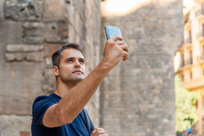 Young man photographing by mobile phone while standing against building