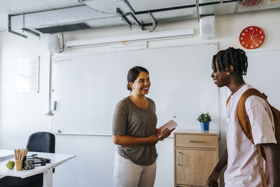 Happy female teacher holding diary while talking with male student in classroom at school