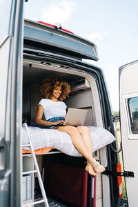 Young african american female traveler with curly hair watching movie on laptop while resting inside camper van during summer holidays