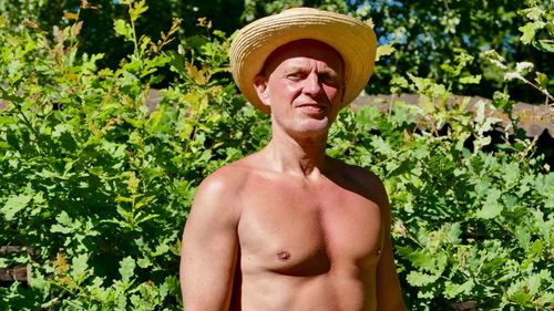 Portrait of shirtless man wearing hat standing against plants