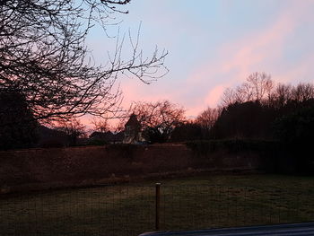 Bare trees on landscape against sky at sunset